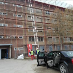 Jankes-Bud Janusz Smolen - Wysoki budynek mieszkalny w trakcie prac na rusztowaniu, długa aluminiowa drabina oparta o ścianę, pracownicy w odblaskowych kamizelkach i kaskach, otwarte drzwi czarnego samochodu, baner reklamowy...