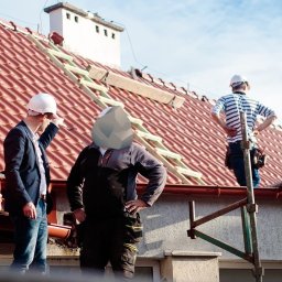 Jankes-Bud Janusz Smolen - Trzech mężczyzn w kaskach na dachu: jeden w garniturze wskazuje palcem, drugi w roboczym ubraniu stoi z rękami na biodrach, trzeci w koszulce w paski stoi na rusztowaniu. Dach pokryty czerwoną...