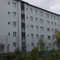 Wielopiętrowy budynek mieszkalny pomalowany na pastelowy odcień zieleni, widoczny zza siatki ogrodzeniowej porośniętej bluszczem.