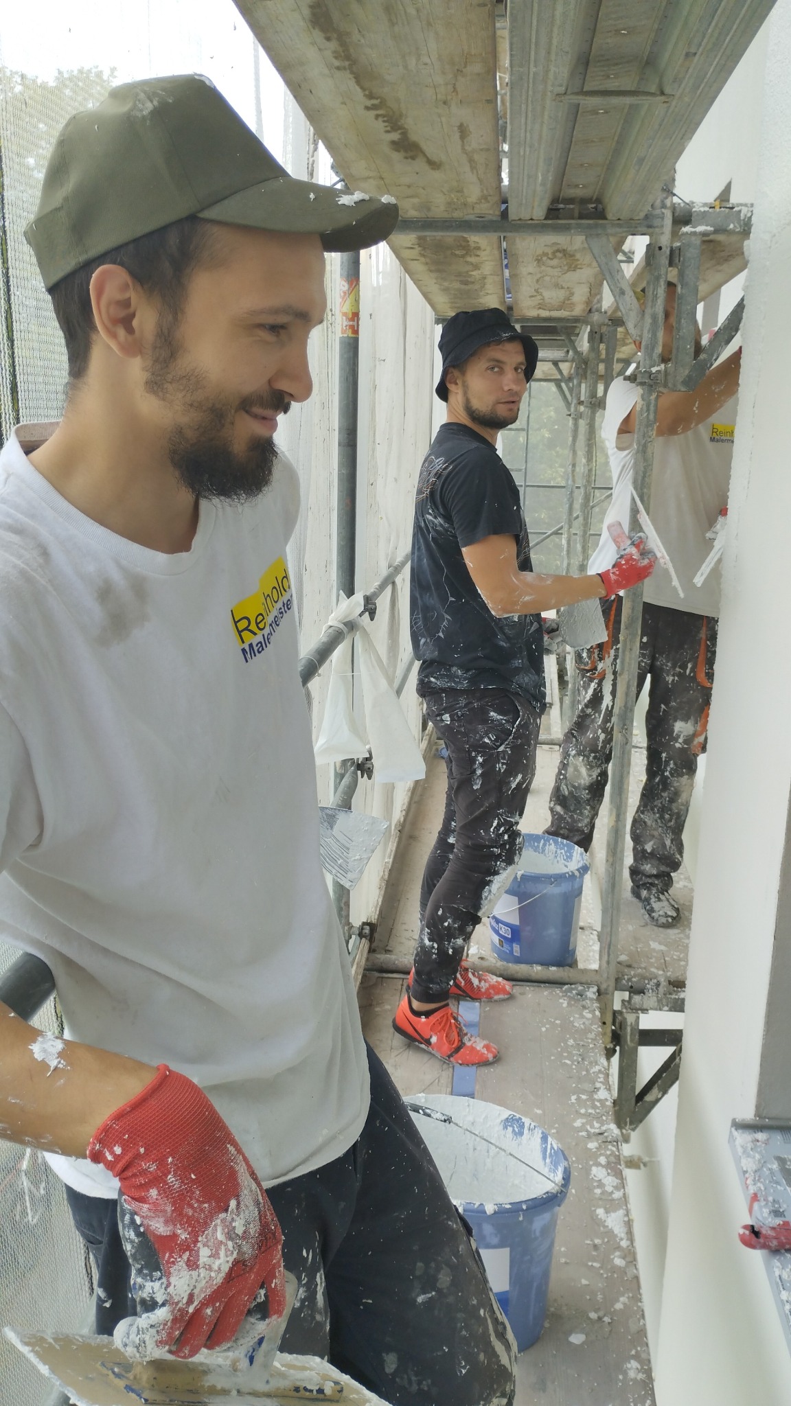 Trzech mężczyzn na rusztowaniu, jeden z brodą w zielonej czapce z daszkiem, uśmiecha się i trzyma szpachelkę z zaprawą, w tle dwaj inni pracownicy wygładzają ścianę, widoczne wiadra z zaprawą...