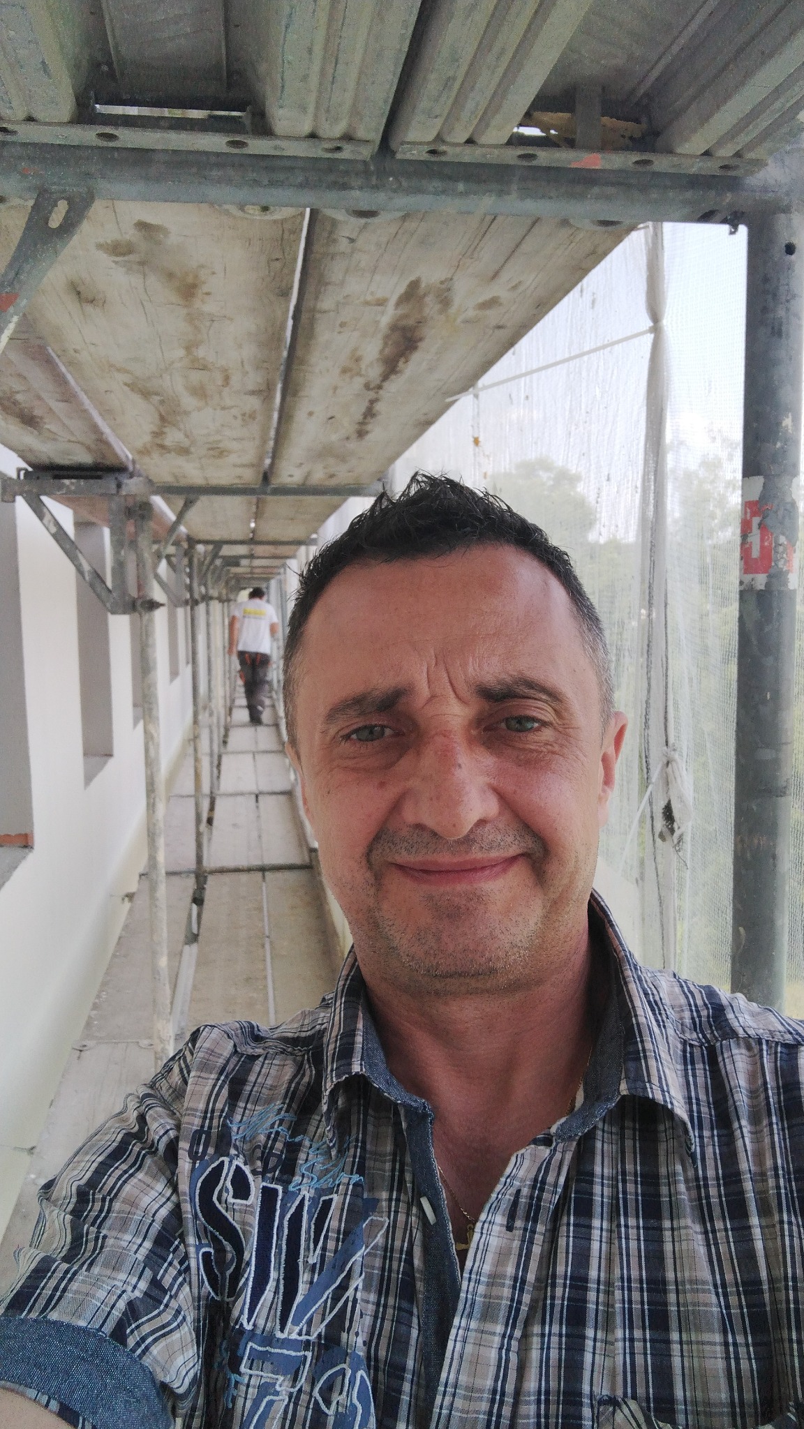 Uśmiechnięty mężczyzna w koszuli w kratę na rusztowaniu, widoczny fragment otynkowanej ściany budynku i inny pracownik w oddali.