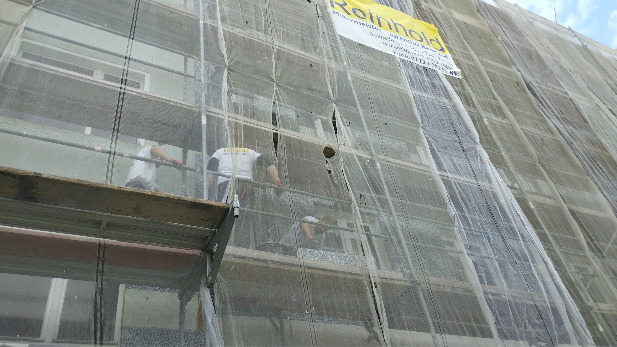 Prace na wysokościach: Robotnicy na rusztowaniu, zabezpieczeni siatką, odnawiają elewację budynku z jasną fasadą i oknami, widoczny baner reklamowy firmy Reinhold.