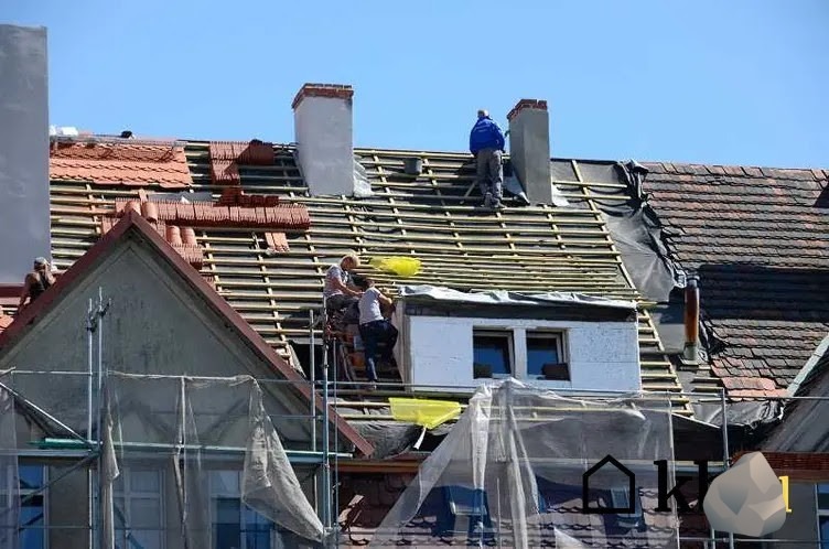 Ekipa dekarzy na dachu podczas wymiany pokrycia, widoczne łatkowanie, kominy i częściowo ułożona dachówka ceramiczna, zabezpieczenia rusztowań w dolnej części kadru.