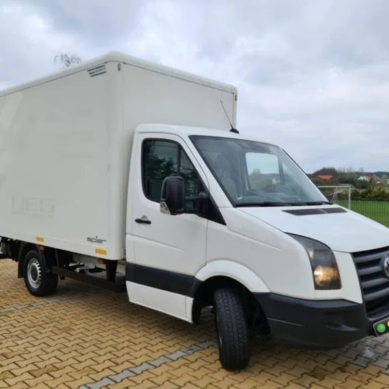 Biały samochód dostawczy Volkswagen Crafter z zabudową kontenerową, zaparkowany na kostce brukowej, widoczny z boku na tle pochmurnego nieba i zieleni.