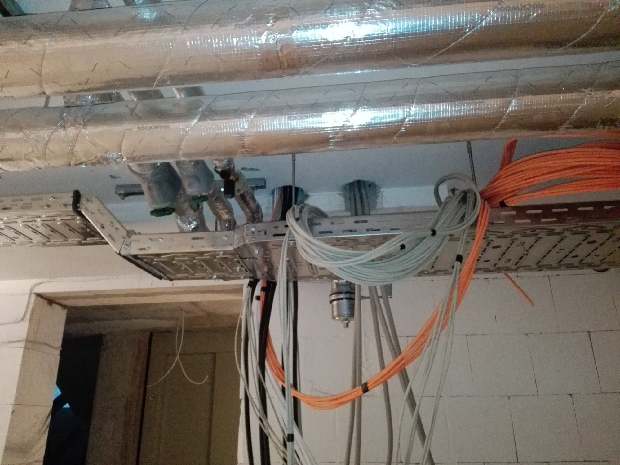 Instalacja przewodów elektrycznych i rur pod sufitem, widoczne kable pomarańczowe i białe, rury izolowane folią aluminiową, metalowe korytka kablowe.