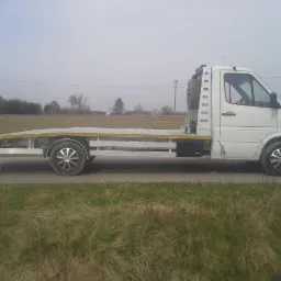 Biała laweta z platformą o kratownicowej strukturze, zaparkowana na poboczu drogi polnej, z zapasowym kołem umieszczonym za kabiną.