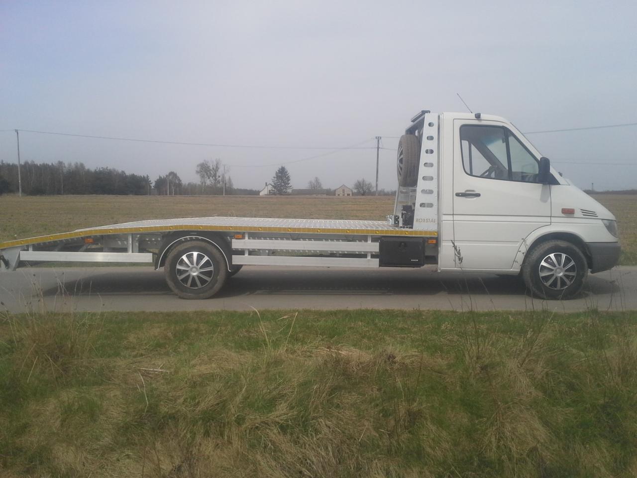 Biała laweta z platformą o kratownicowej strukturze, zaparkowana na poboczu drogi polnej, z zapasowym kołem umieszczonym za kabiną.