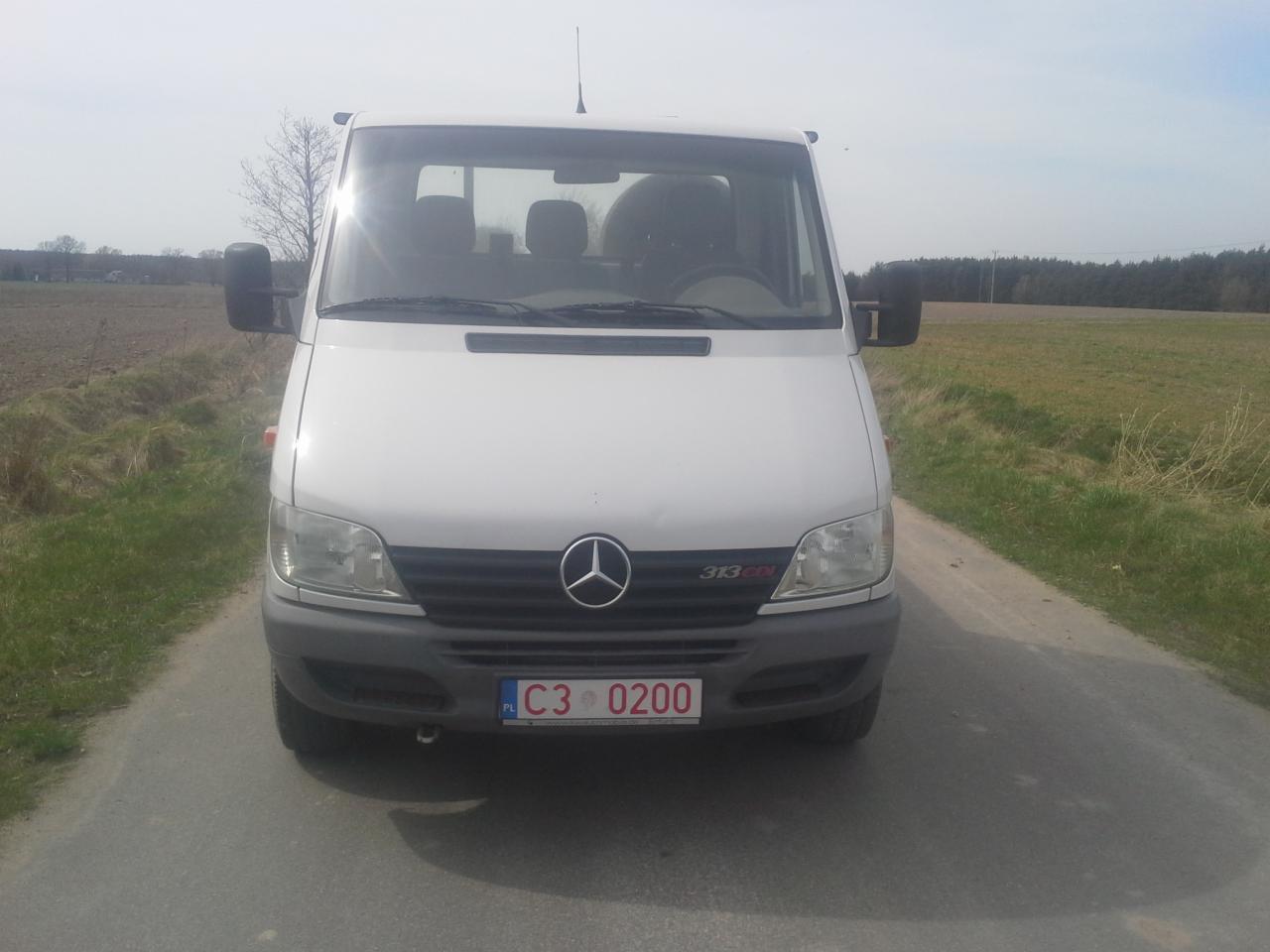 Przód białego busa Mercedes-Benz Sprinter 313 CDI z polską tablicą rejestracyjną C3 0200 na asfaltowej drodze w wiejskim krajobrazie.