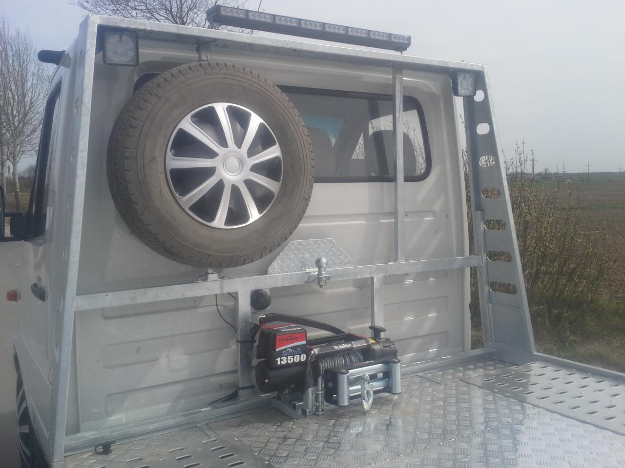 Biała platforma samochodu z zamontowaną wyciągarką Titanium 13500, kołem zapasowym i oświetleniem LED na metalowej konstrukcji, widok z tyłu na tle pola.