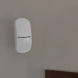 Biały czujnik ruchu alarmowego zamontowany na jasnej ścianie, widoczny fragment ramy obrazu po lewej i fragment drewnianej listwy przypodłogowej po prawej.