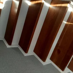 Nowoczesne schody z drewna i białych elementów, oświetlone naturalnym światłem, z widoczną strukturą drewna i szarym dywanem na podłodze. Ujęcie z dołu.