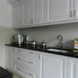 Białe, klasyczne szafki kuchenne z delikatnymi złotymi uchwytami, czarnym blatem i zlewozmywakiem, widoczne garnki i ekspres do kawy.