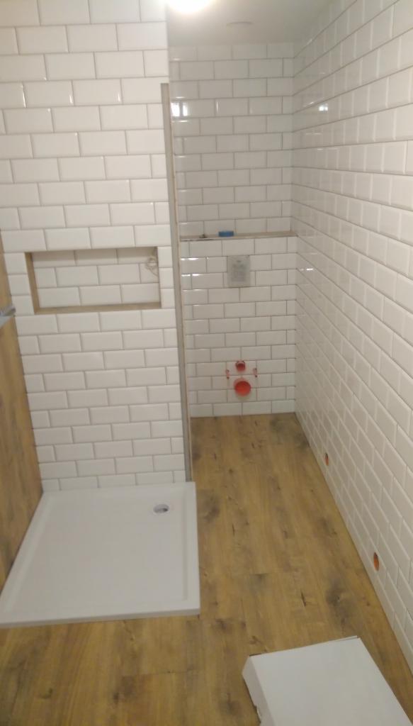 Nowoczesna łazienka w trakcie wykańczania, białe kafelki ułożone w cegiełkę, drewnopodobna podłoga, brodzik i wnęka w ścianie