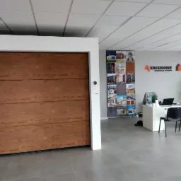 Wystawowa, segmentowa brama z paneli imitujących drewno, widoczna w salonie sprzedaży z biurkiem i ścianą z logotypem firmy w tle.