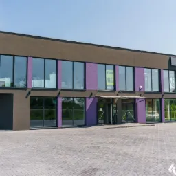 Nowoczesny budynek biurowy z dużymi oknami i wejściem, pomalowany w odcieniach szarości i fioletu, z widocznym logo firmy na elewacji.
