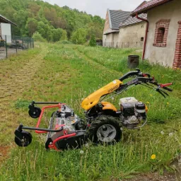 Żółto-czarno-czerwona kosiarka listwowa XB40 na wysokiej trawie z dmuchawcami, tło stanowią budynki gospodarcze i las.