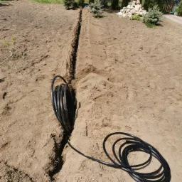 Wykopany rów w ziemi z czarnym wężem do nawadniania w ogrodzie, w tle widoczne drzewa iglaste i budynek gospodarczy.