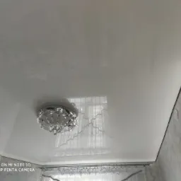 Biały, błyszczący sufit napinany z ozdobnym żyrandolem, widoczne refleksy światła i fragment ściany o szarej fakturze.