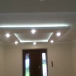 Nowoczesny sufit podwieszany z oświetleniem LED punktowym i listwami dekoracyjnymi, widok na fragment ściany z dekoracyjną fakturą kamienia i drzwi wejściowe.