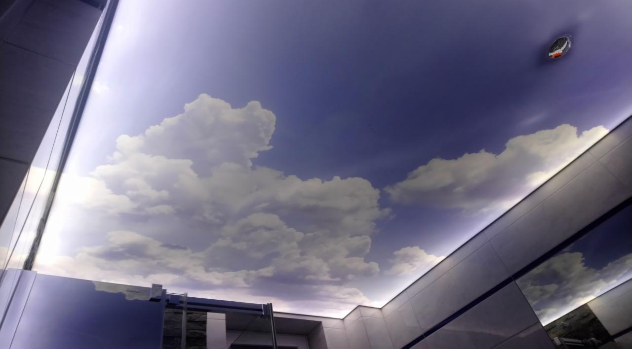 Sufit w łazience z fototapetą przedstawiającą błękitne niebo z chmurami, oświetlony listwą LED, z widocznym czujnikiem dymu.