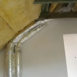 Dwa przewody wentylacyjne owinięte folią aluminiową, biegnące wzdłuż ściany i pod sufitem z izolacją z wełny mineralnej, widoczne elementy drewnianej konstrukcji.