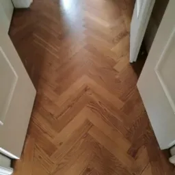 Ułożony parkiet w jodełkę, widoczne drzwi po bokach, naturalne drewno z wyraźnym usłojeniem.