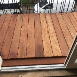 Nowoczesna drewniana antresola z desek w odcieniach brązu, widok z wnętrza przez otwarte drzwi balkonowe, z barierką i białymi furgonetkami w tle.