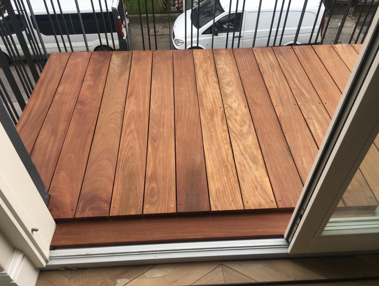 Nowoczesna drewniana antresola z desek w odcieniach brązu, widok z wnętrza przez otwarte drzwi balkonowe, z barierką i białymi furgonetkami w tle.