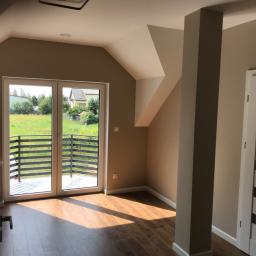 MARBUD - MARIAN SKOTNIK - Jasne wnętrze poddasza z drewnianą podłogą, widokiem na zieleń z balkonu i ścianami w odcieniach beżu, z widoczną kolumną i częścią białych drzwi z szarymi wstawkami.