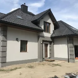 Nowoczesny dom jednorodzinny z szarą elewacją i ciemną dachówką, widoczne detale architektoniczne, takie jak boniowanie narożników i obramowania okien, teren wokół domu w trakcie prac wykończeniowych.