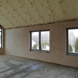 MARBUD - MARIAN SKOTNIK - Surowe wnętrze z trzema oknami, ściany pokryte świeżym tynkiem, izolacja piankowa na skośnym suficie, betonowa podłoga w trakcie prac.
