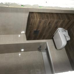 MARBUD - MARIAN SKOTNIK - Wykończona łazienka z jasnymi płytkami podłogowymi i częściowo obłożoną drewnopodobnymi płytkami ścianą, widoczna toaleta i wanna z baterią.