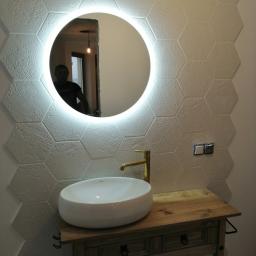 MARBUD - MARIAN SKOTNIK - Białe heksagonalne płytki ścienne z subtelnym wzorem, okrągłe lustro z podświetleniem LED, złota bateria umywalkowa i owalna umywalka nablatowa na drewnianej szafce z szufladami.