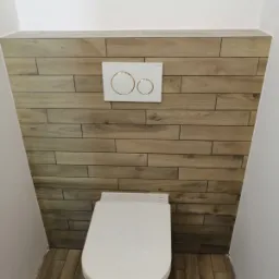 Podwieszana toaleta z białą deską i przyciskiem spłukującym na ścianie wyłożonej płytkami imitującymi drewno, wąskie pomieszczenie.