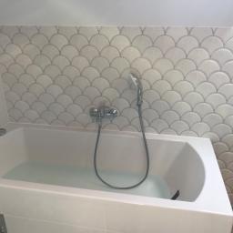 MARBUD - MARIAN SKOTNIK - Wanna wypełniona wodą, z baterią i słuchawką prysznicową na tle ściany wyłożonej białymi płytkami w kształcie wachlarzy.