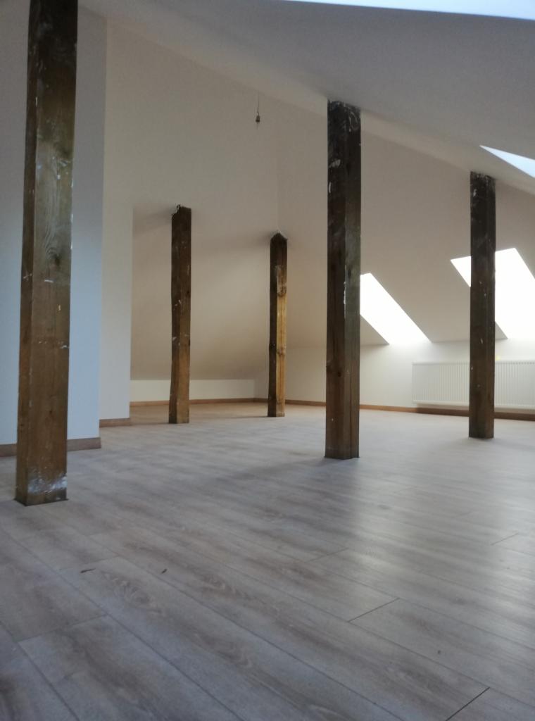 Podłoga z paneli w jasnym odcieniu drewna, drewniane słupy konstrukcyjne w surowym stanie, białe ściany i skosy poddasza z oknami dachowymi, grzejnik na ścianie.