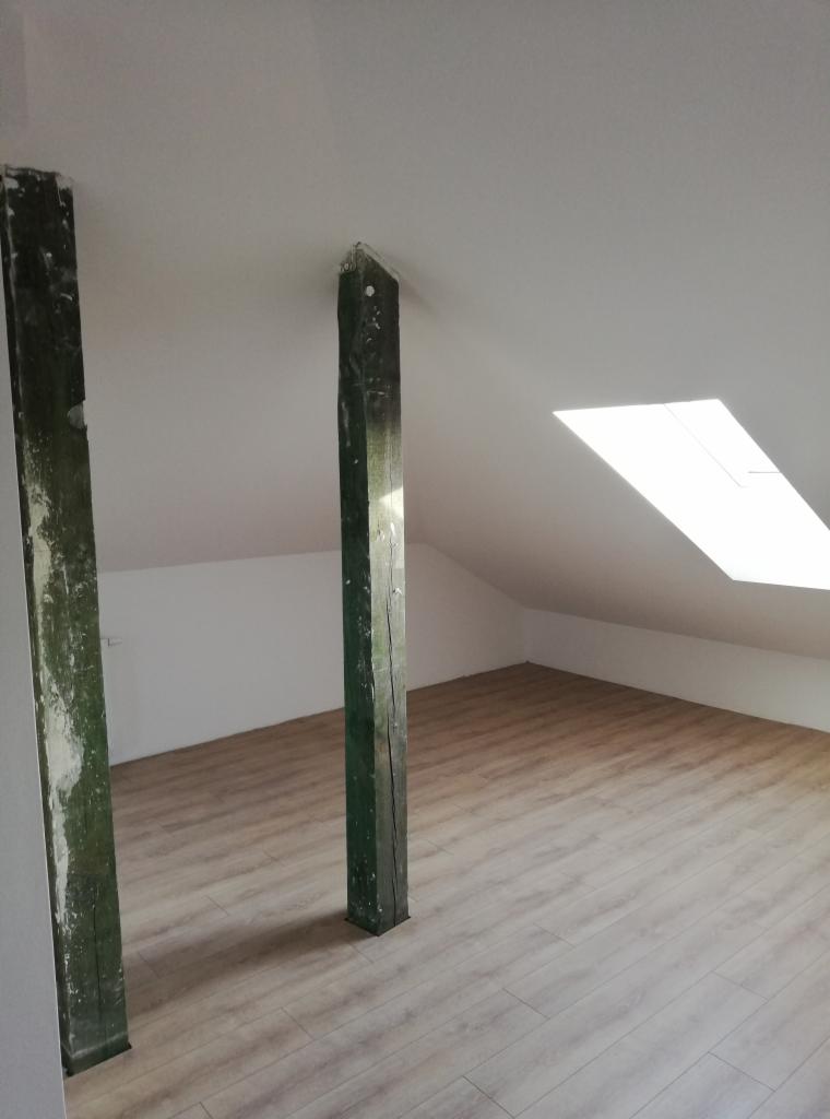 Poddasze po remoncie z nową podłogą panelową i odnowionymi, zielonymi, drewnianymi słupami konstrukcyjnymi, widoczne okno dachowe.