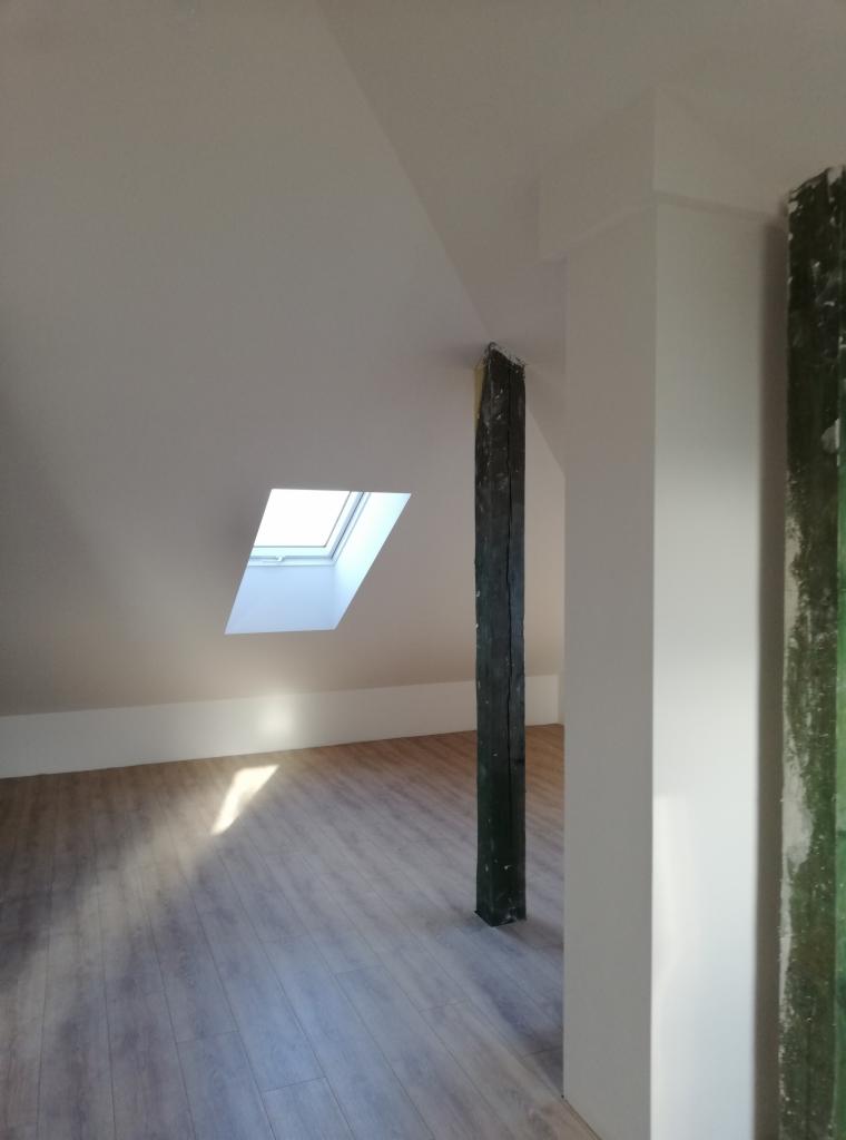Poddasze po remoncie z nowymi panelami podłogowymi, oknem dachowym i odnowioną drewnianą belką, widoczna ściana po malowaniu.