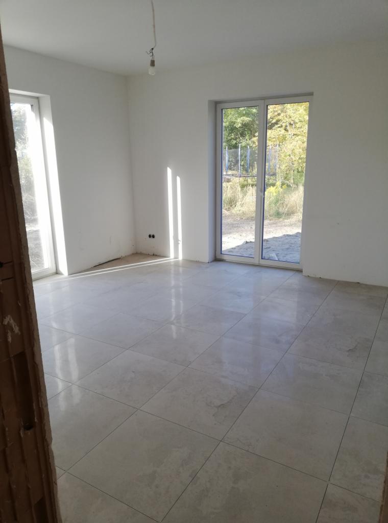 Jasne, kwadratowe płytki podłogowe z delikatnym wzorem w nowym, jeszcze niewykończonym pokoju z dużym oknem balkonowym i oknem z widokiem na zieleń.