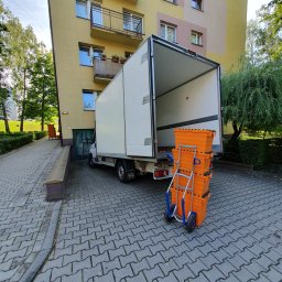 Rentic - Otwarta paka białego samochodu dostawczego zaparkowanego przed blokiem, obok wózek transportowy z trzema pomarańczowymi pojemnikami.