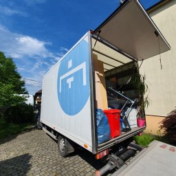 Rentic - Ciężarówka z otwartą przestrzenią ładunkową, wypełniona kartonami, rośliną doniczkową w różowej doniczce, czerwonym pojemnikiem i niebieskim workiem, widziana z tyłu na brukowanej drodze...