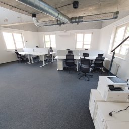 Rentic - Jasne, przestronne biuro z rzędami białych biurek oddzielonych przegrodami, czarnymi krzesłami obrotowymi i widocznymi instalacjami wentylacyjnymi pod betonowym sufitem.