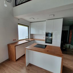 Rentic - Jasna kuchnia z drewnianymi blatami i wyspą kuchenną, okno z osłoną, widoczne instalacje elektryczne, wnętrze w trakcie wykańczania.