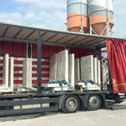 Czerwona ciężarówka z otwartą naczepą załadowana prefabrykatami betonowymi zabezpieczonymi niebieskimi pasami transportowymi, na tle silosy do betonu.