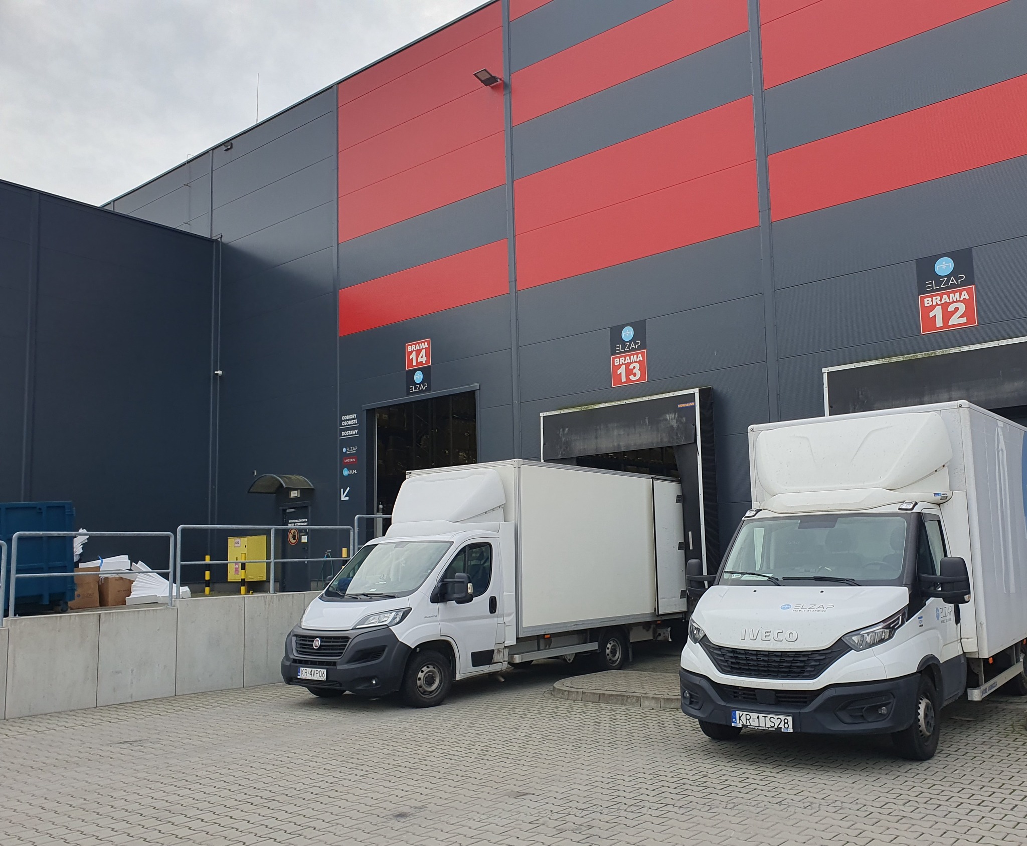 Dwa białe busy dostawcze Iveco i Fiat zaparkowane przed rampami magazynu z czerwono-szarymi pasami, widoczne numery bram 12, 13 i 14 oraz oznaczenia Elzap.