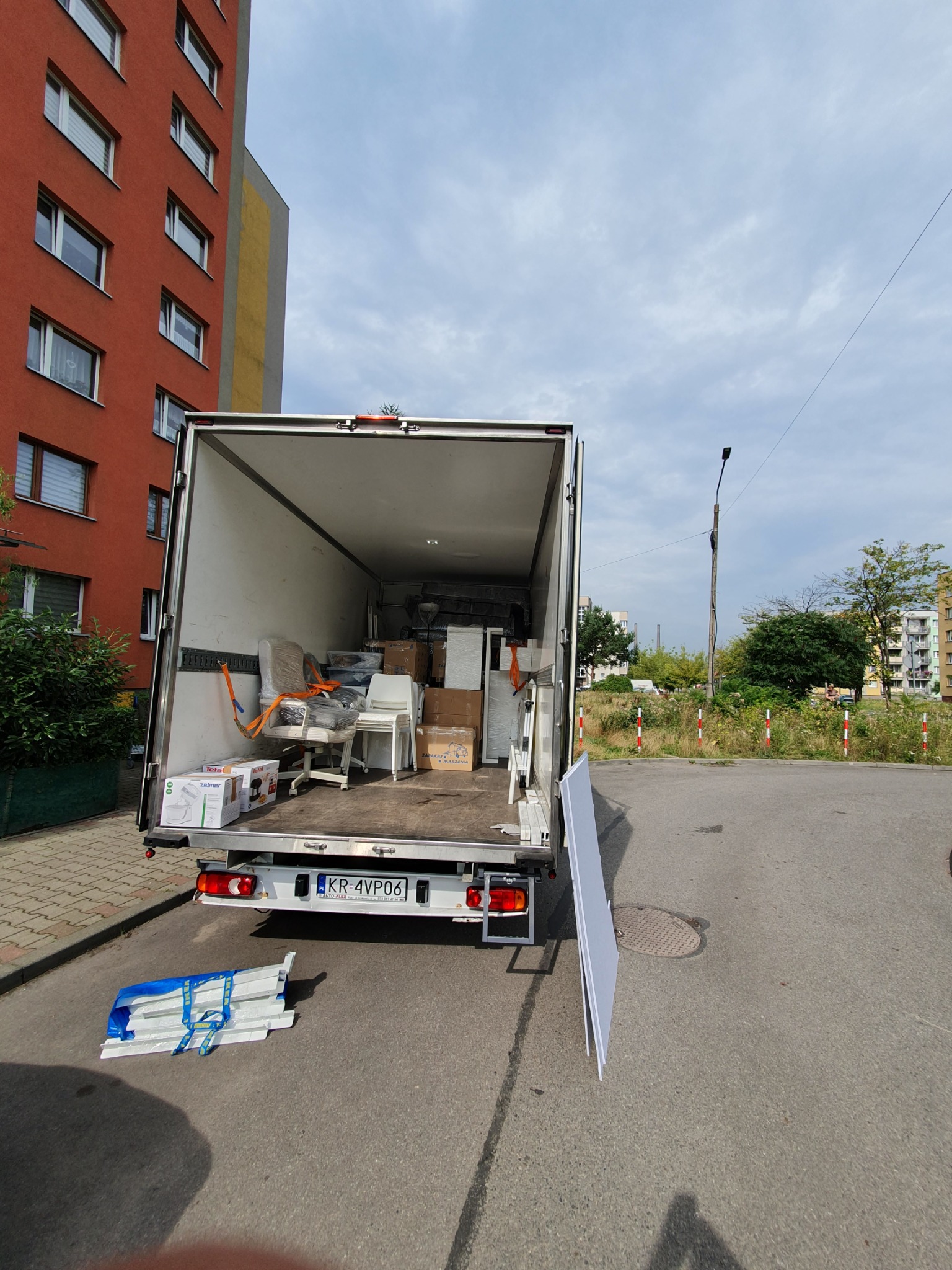 Otwarta paka samochodu dostawczego wypełniona meblami i kartonami, widoczne krzesła biurowe owinięte folią, kartony z mikserami, na zewnątrz leżą elementy meblowe zabezpieczone niebieskimi pasami...