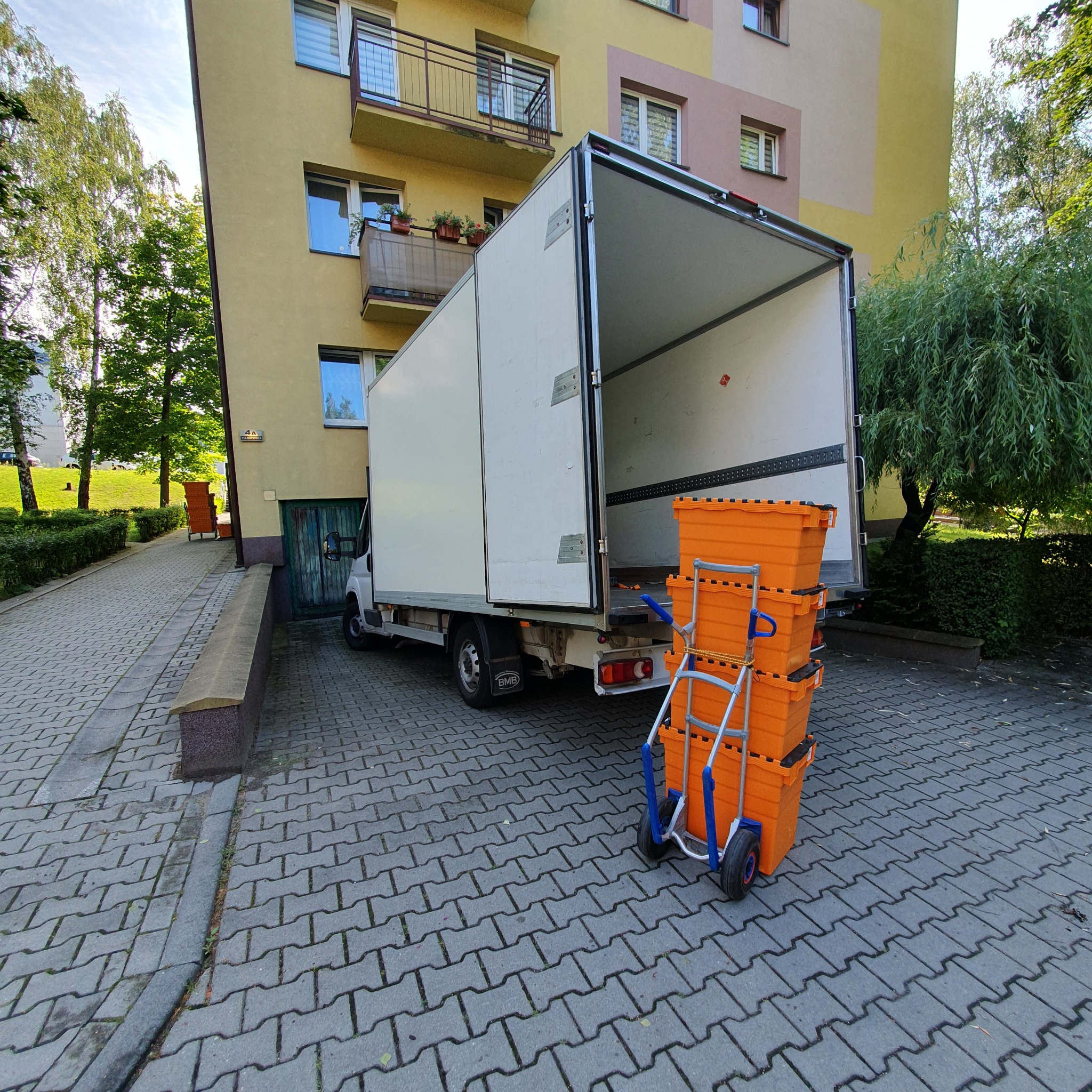 Otwarta paka białego samochodu dostawczego zaparkowanego przed blokiem, obok wózek transportowy z trzema pomarańczowymi pojemnikami.