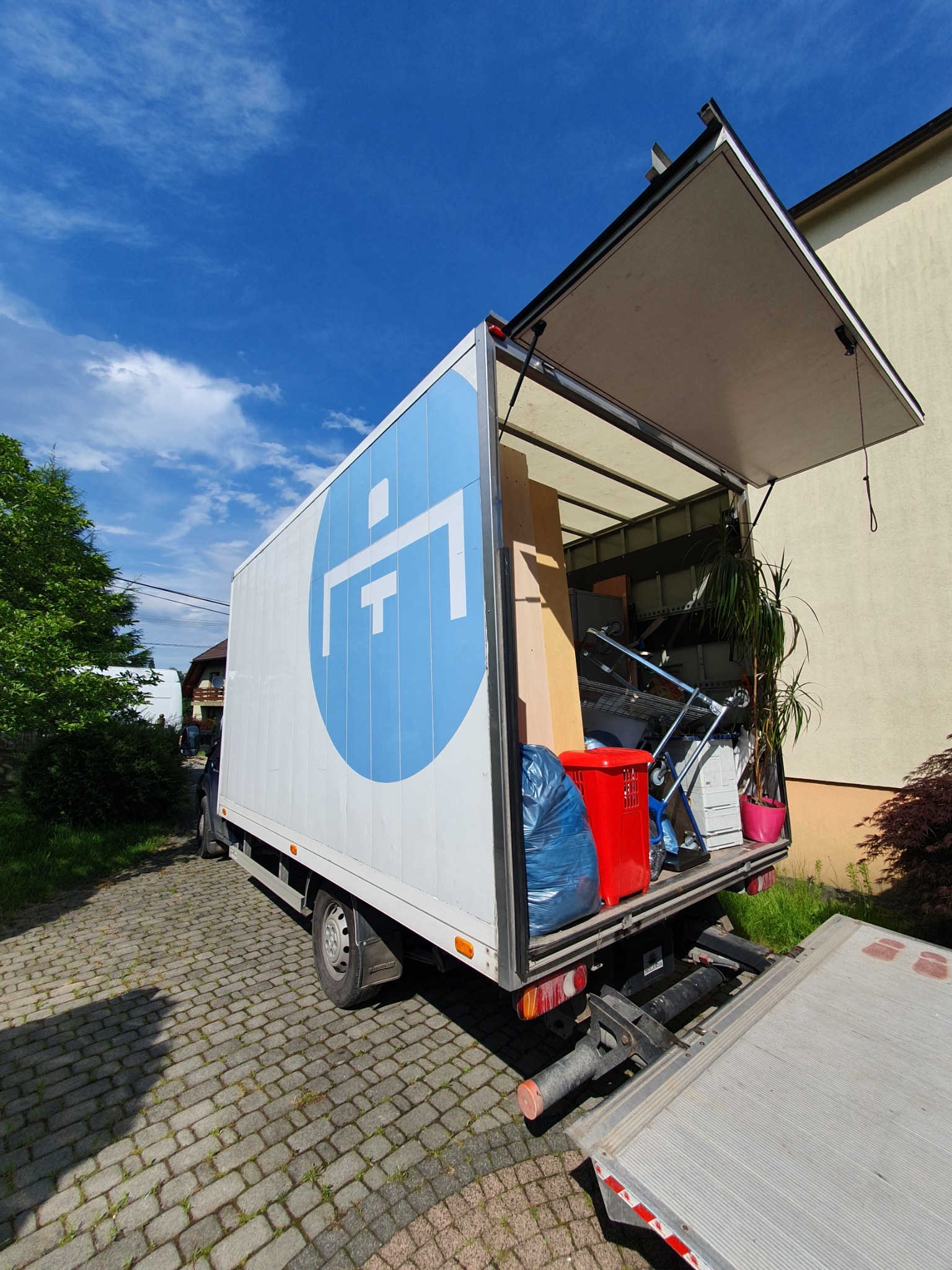 Ciężarówka z otwartą przestrzenią ładunkową, wypełniona kartonami, rośliną doniczkową w różowej doniczce, czerwonym pojemnikiem i niebieskim workiem, widziana z tyłu na brukowanej drodze...