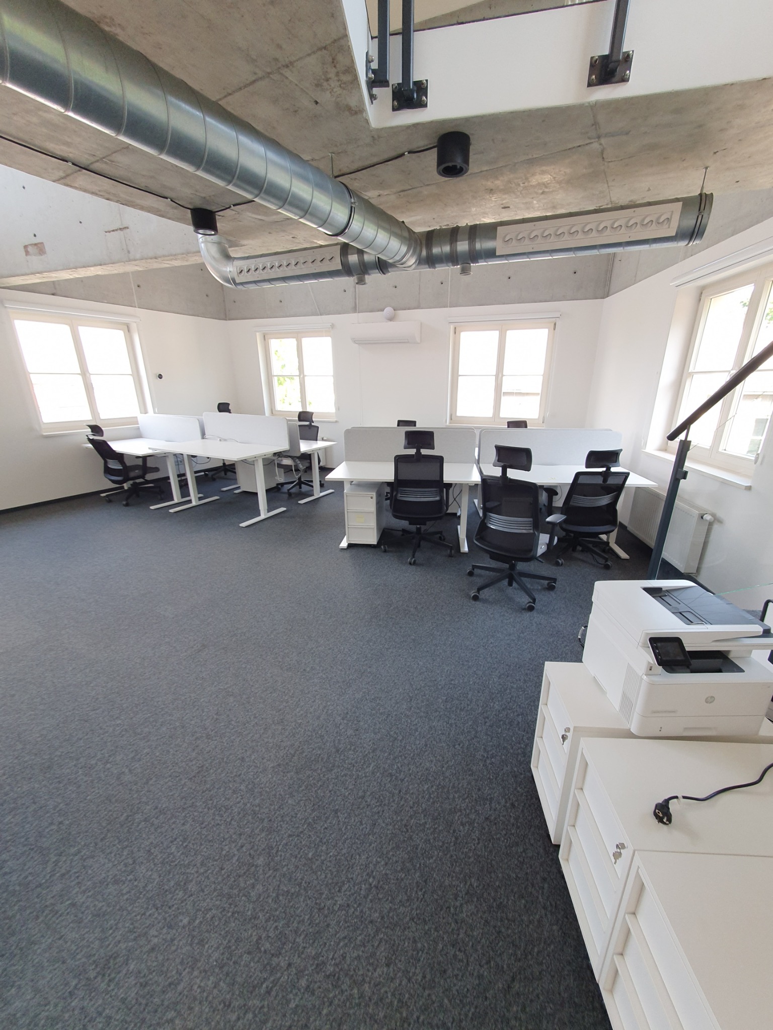 Jasne, przestronne biuro z rzędami białych biurek oddzielonych przegrodami, czarnymi krzesłami obrotowymi i widocznymi instalacjami wentylacyjnymi pod betonowym sufitem.