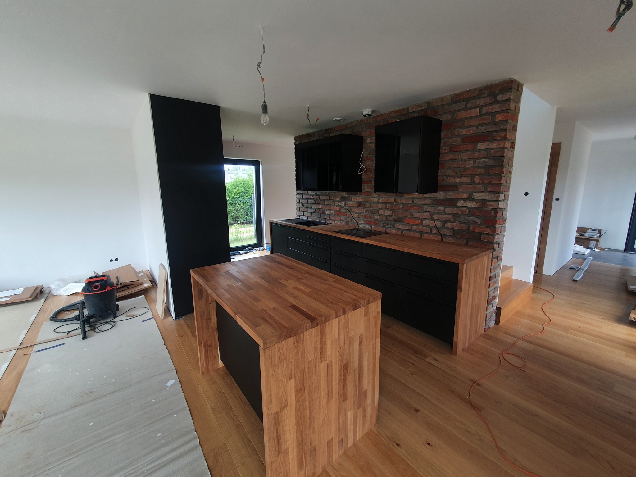 Wnętrze kuchni w trakcie montażu: surowa cegła na ścianie, drewniany blat, czarne szafki, wyspa kuchenna z drewnianym wykończeniem, widoczny odkurzacz przemysłowy i kartony zabezpieczające podłogę.